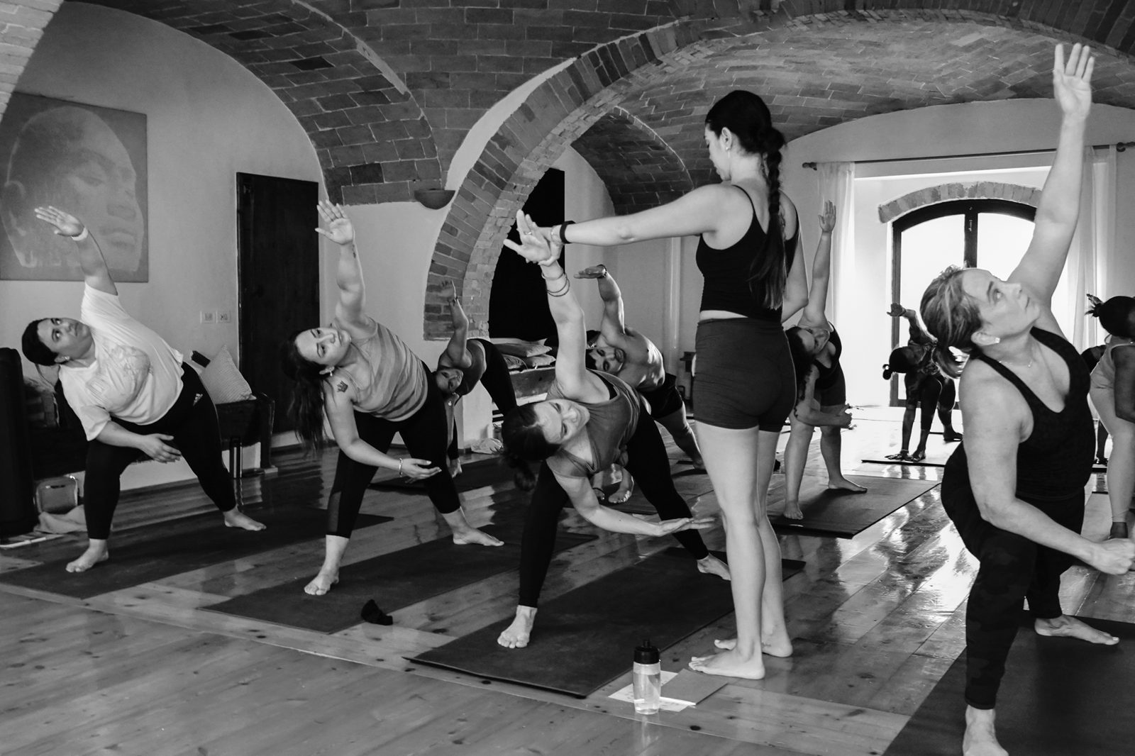 Black and white photo of Irene del Valle leading a group Ashtanga class in a vaulted brick studio